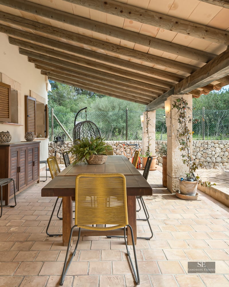 Covered terrace with wooden table, chairs, stone walls, and beamed ceiling overlooking the countryside.