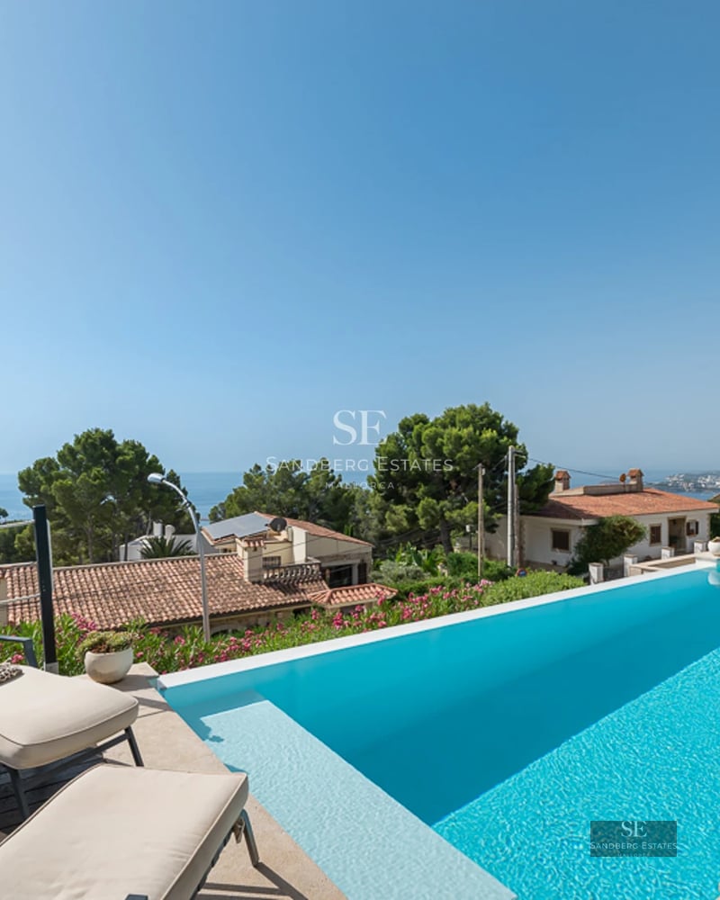 View of a luxury villa with infinity pool, sun loungers, and lush garden. Modern design featuring large windows.