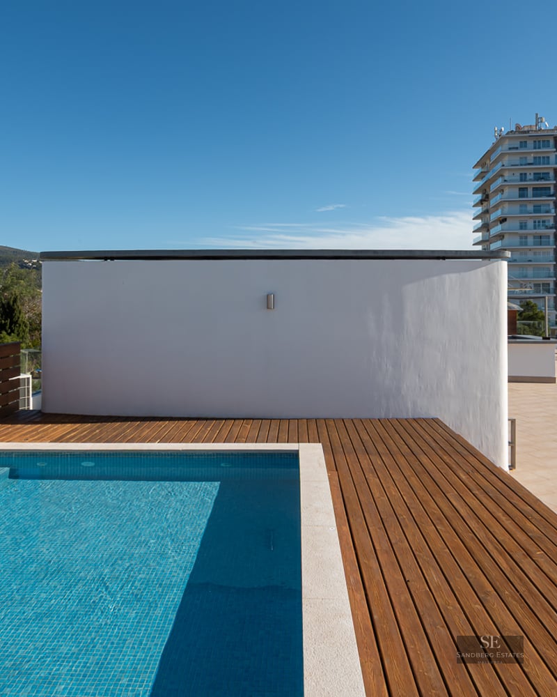 Piscine moderne sur le toit avec terrasse en bois, vue sur la mer et les montagnes sous un ciel bleu clair.