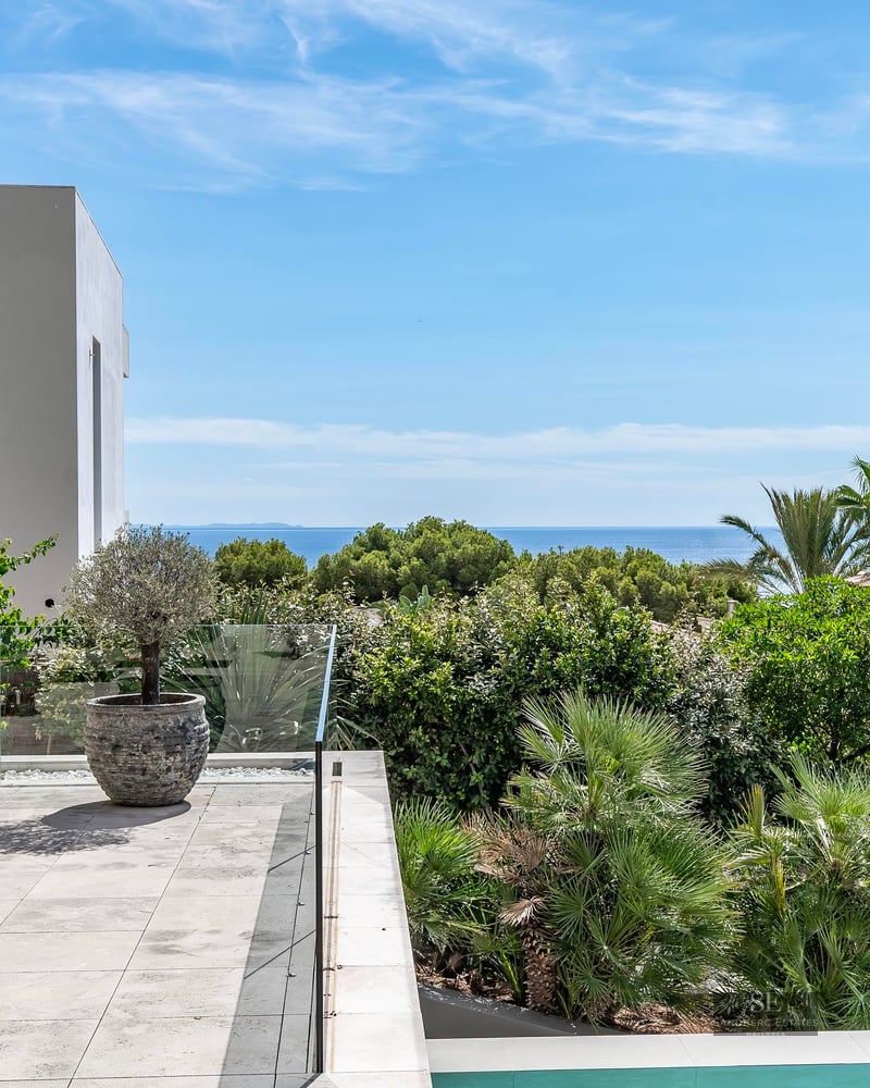 Modern stone terrace with glass railing, sun loungers, and lush green plants overlooking the blue Mediterranean Sea.