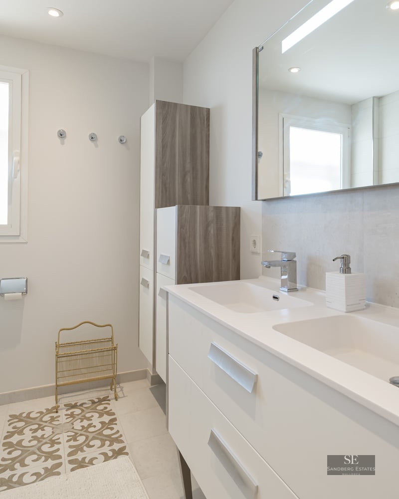 Bright modern bathroom with a double white vanity, large mirror, wall-hung toilet, and natural window light.