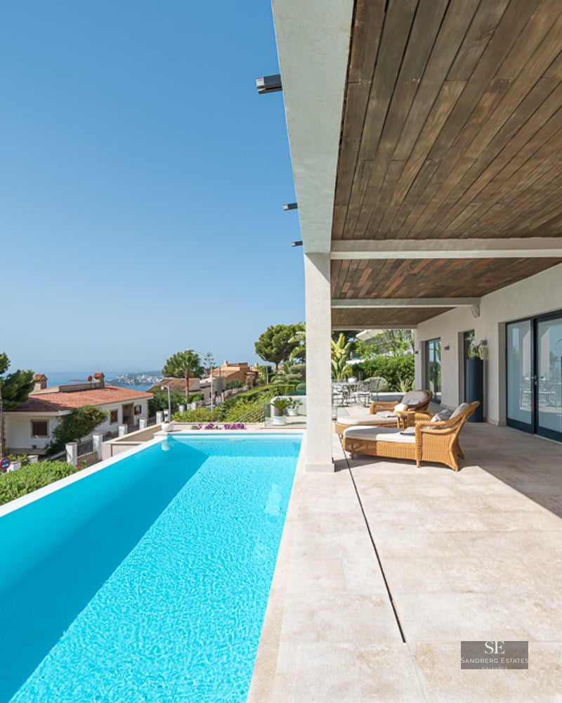 A bright terrace with a turquoise infinity pool, wicker loungers, and a wooden ceiling overlooking a coastal landscape.