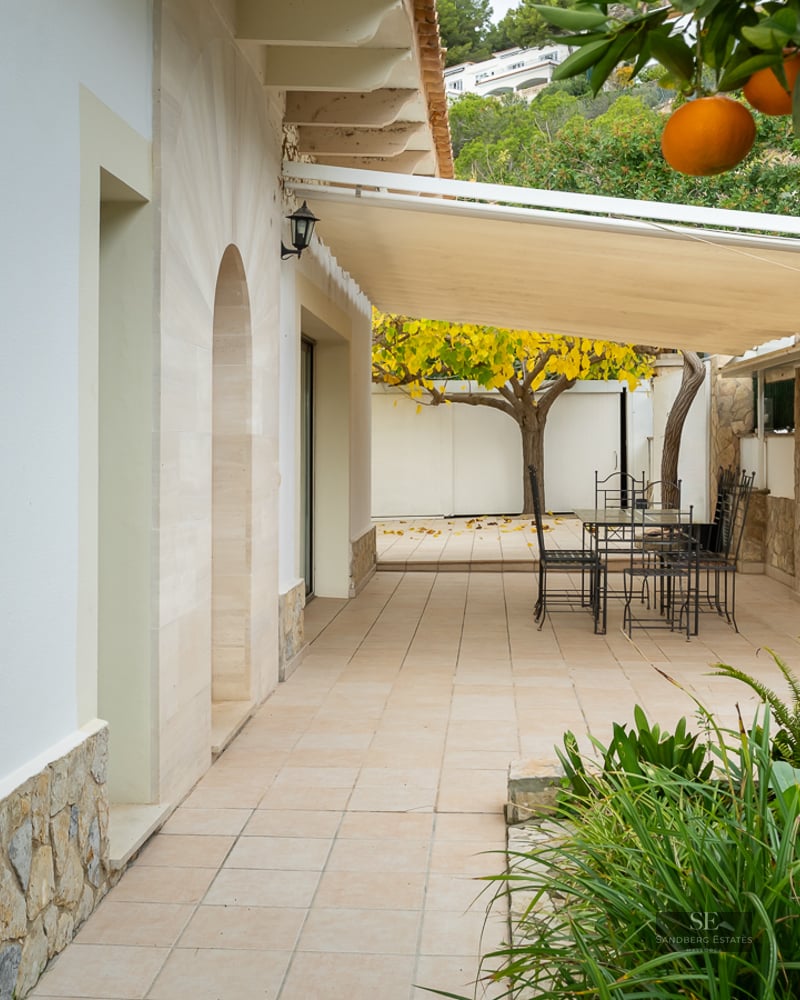 A sun-shaded outdoor terrace with a dining table, black metal chairs, stone walls, and a lush orange tree.