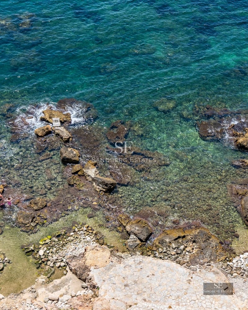 Exterior of modern villa featuring infinity pool, sun loungers, and panoramic Mediterranean sea views. Minimalist architecture and manicured landscaping.
