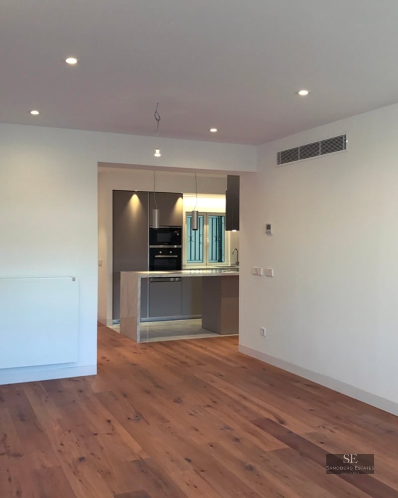 Spacious empty living room with hardwood floors leading to a modern grey kitchen with recessed ceiling lights.