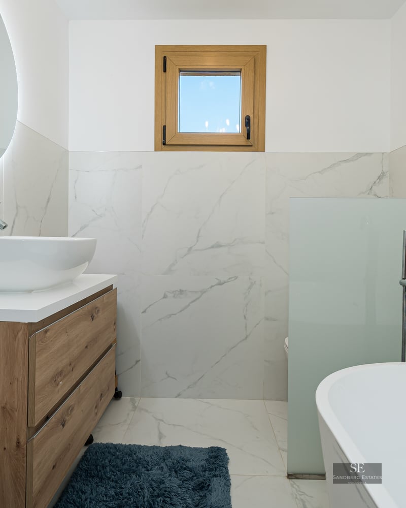 Modern bathroom with marble walls, wood vanity, vessel sink, circular backlit mirror, and a white bathtub.