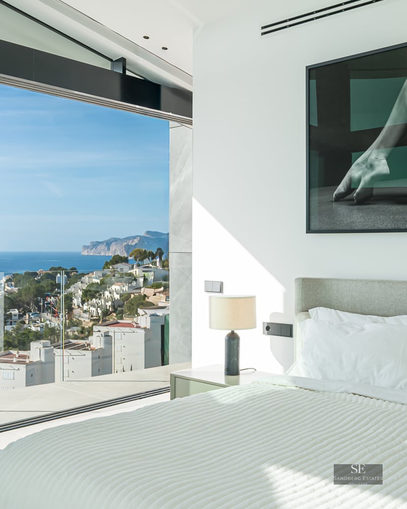 Modern bedroom with large glass windows showing a panoramic view of the sea and coastline.
