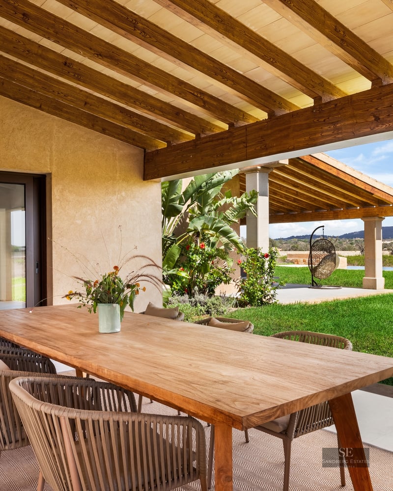 Covered terrace with a large wooden dining table and rope chairs overlooking a bright green lawn and distant hills.