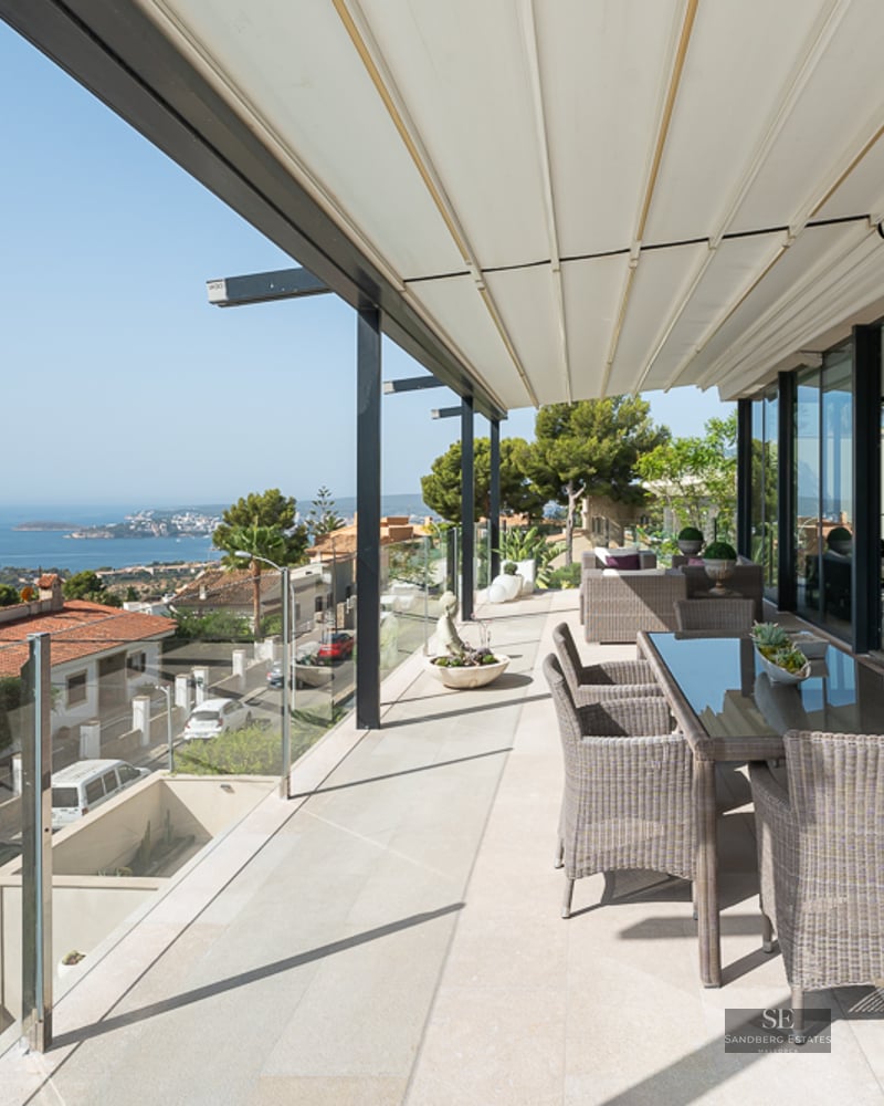 Spacious modern terrace with wicker dining furniture, glass railings, and expansive views of the ocean and coastal town.