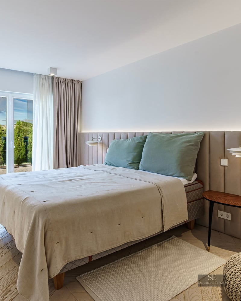 A bright modern bedroom featuring a large bed with a beige upholstered wall headboard and light wood chevron floors.