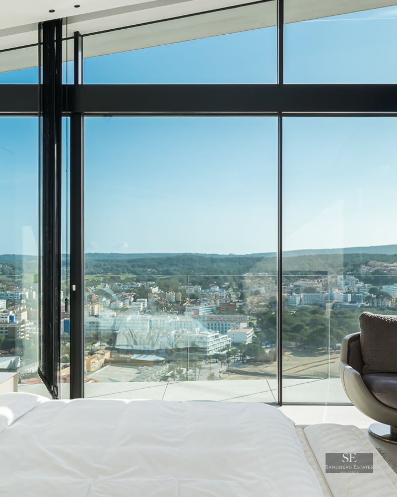 Modern master bedroom featuring floor-to-ceiling windows with a sweeping city view and a designer armchair.