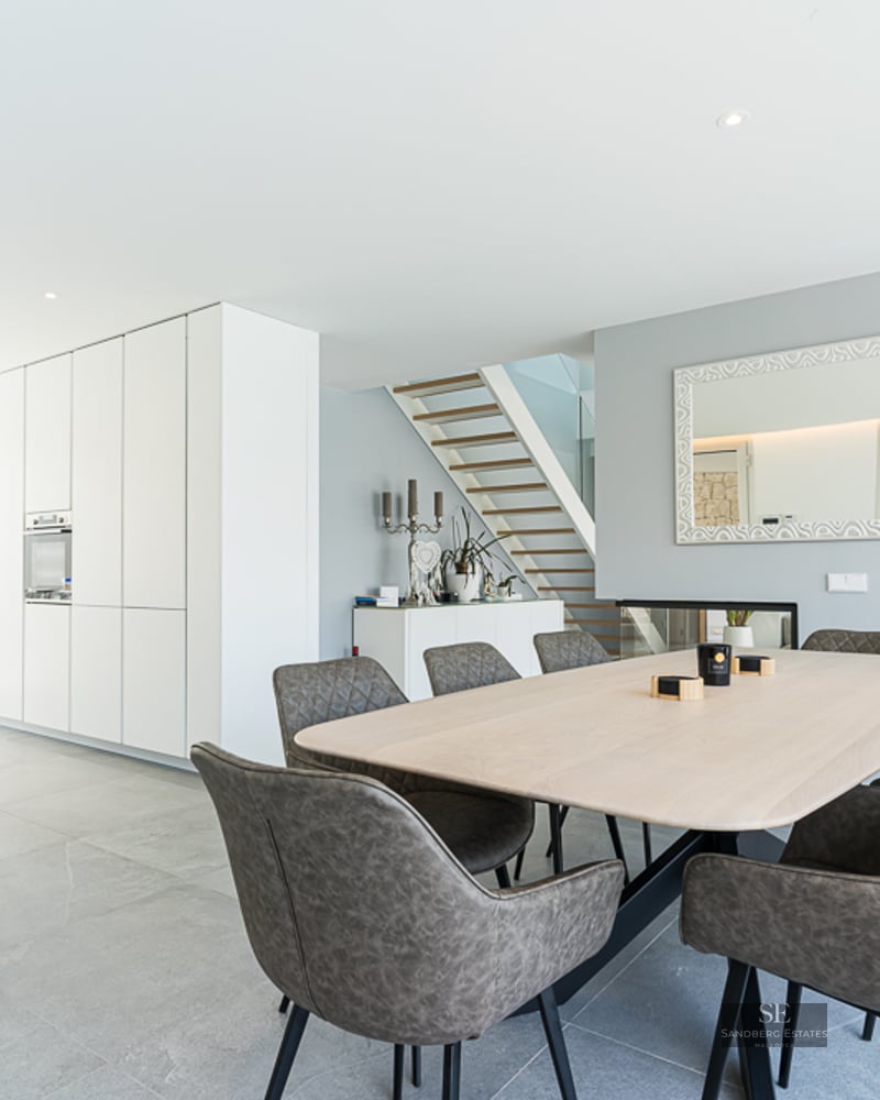 Bright dining area featuring a light wood table, grey chairs, and a minimalist white kitchen in the background.