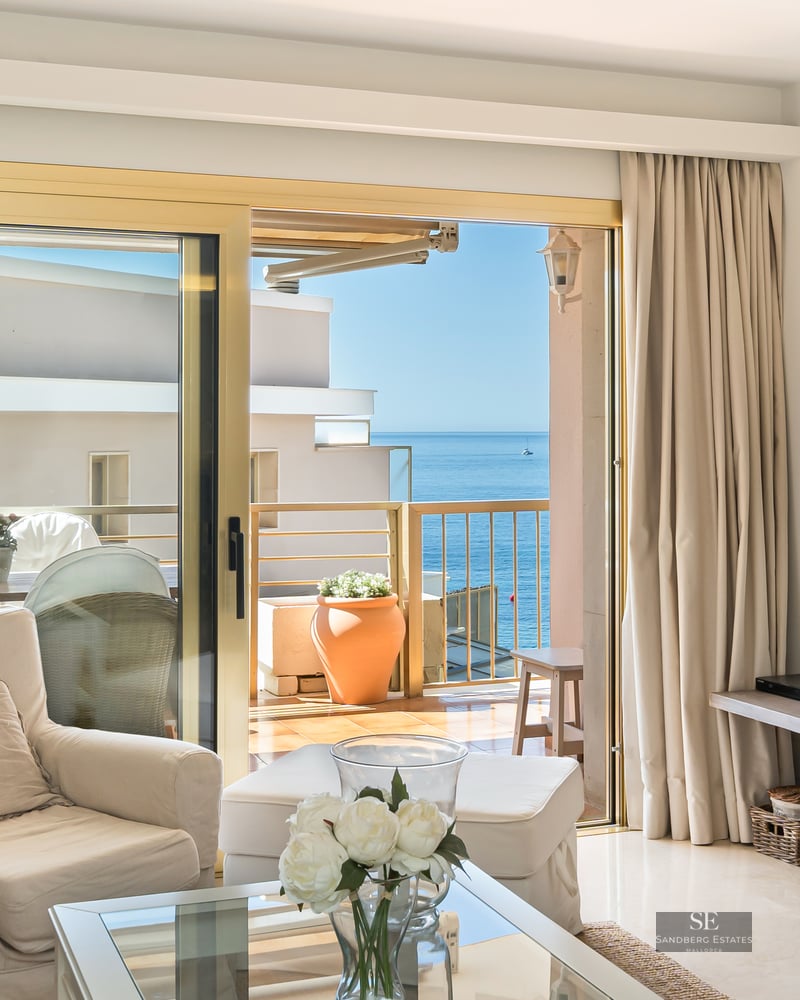 Elegant living room with beige armchair and glass table opening to a terrace with a panoramic Mediterranean sea view.