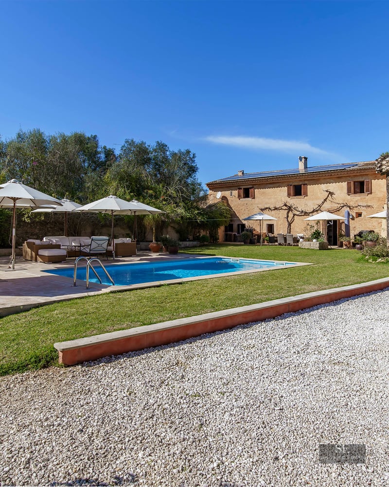 View of a large stone villa with a blue swimming pool, white umbrellas, and garden under a clear sky.