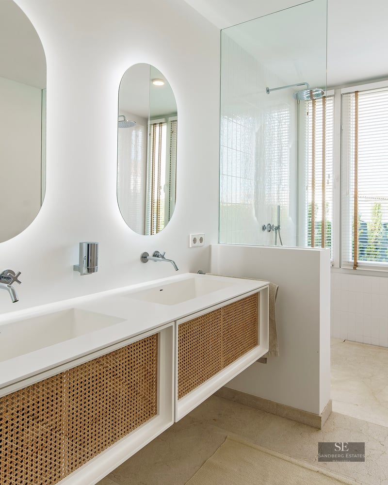 Modern master bathroom featuring a white double vanity with rattan doors, oval backlit mirrors, and a glass-enclosed shower.