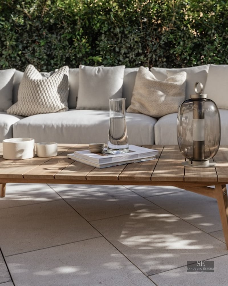 Outdoor sofa with beige cushions and a wooden coffee table on a stone patio against a green hedge.
