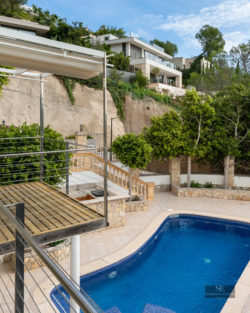 High-angle view of a blue swimming pool, wooden deck, and tiled patio set against a lush green hillside.