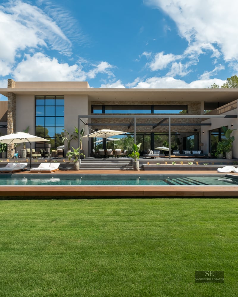 Contemporary villa featuring a lap pool, wood decking, green lawn, and stone facade under a bright blue sky.