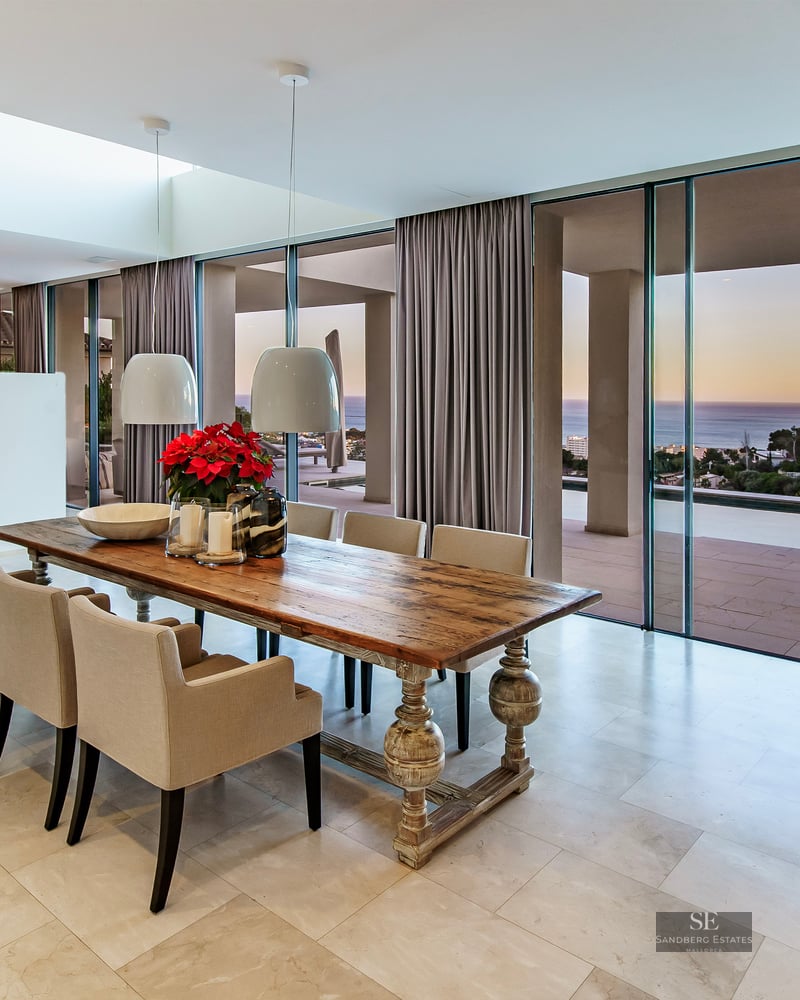 Elegant dining area featuring a long wooden table and floor-to-ceiling windows overlooking the ocean at sunset.