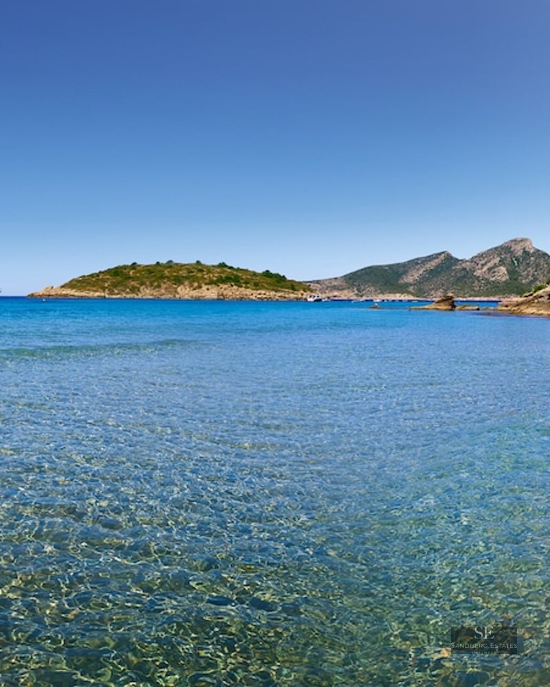 View of a crystal clear turquoise bay with a white villa on a cliff and mountains in the distance.
