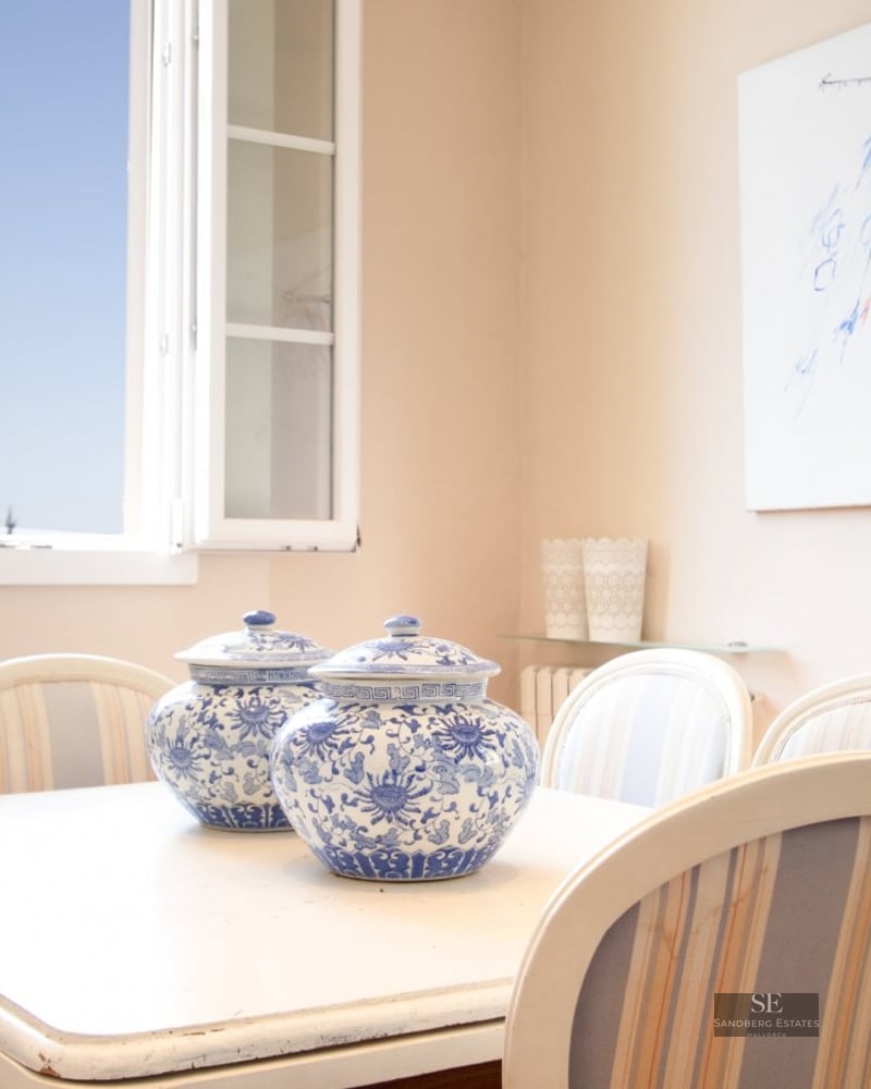 Bright dining area with white table featuring two blue and white porcelain jars and an open window with blue sky view.
