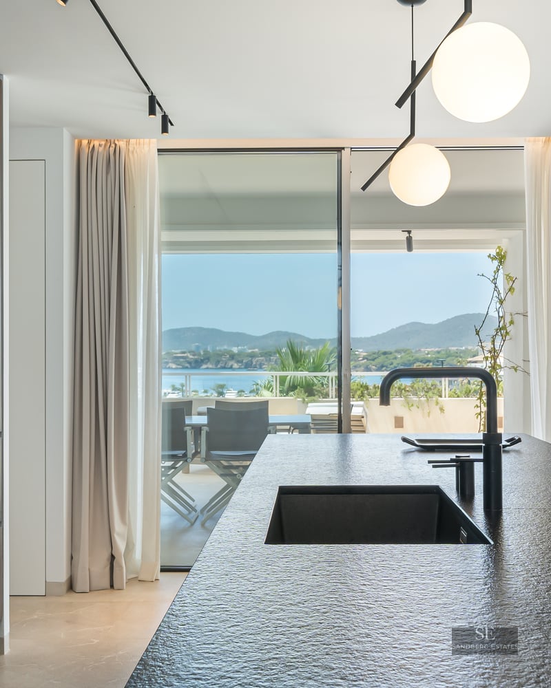 Modern kitchen with a dark stone island and black sink looking out to a sunlit terrace and sea view.