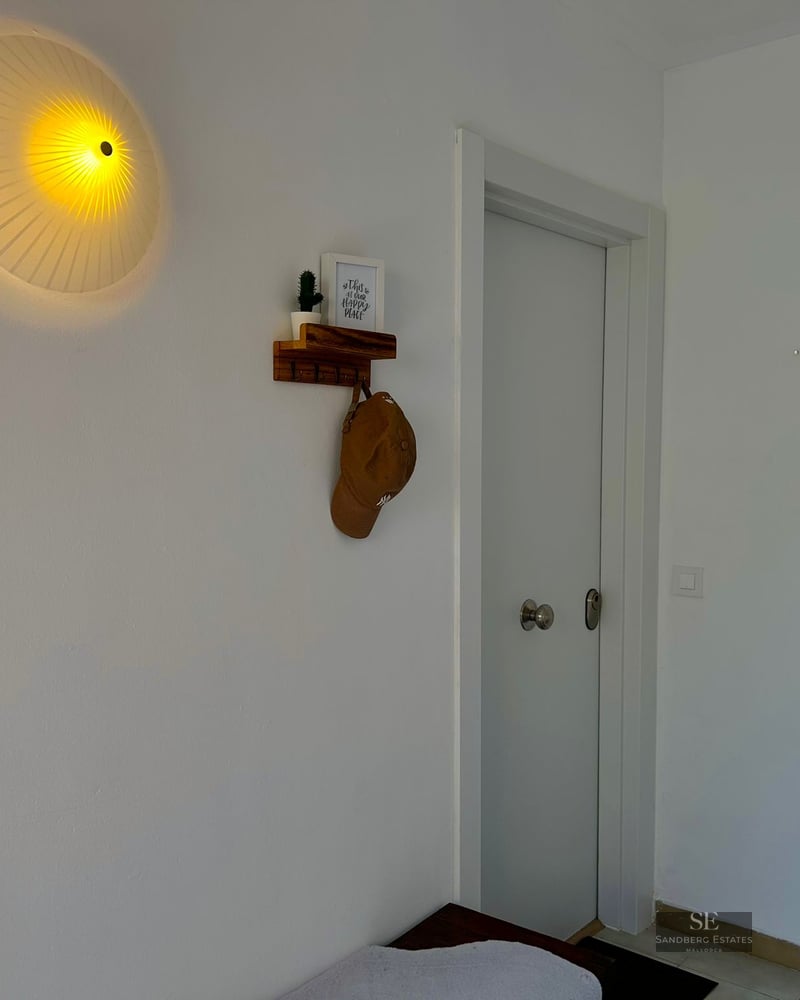 A white hallway featuring a round pleated wall lamp, a small wooden shelf with a hat, and a white door.
