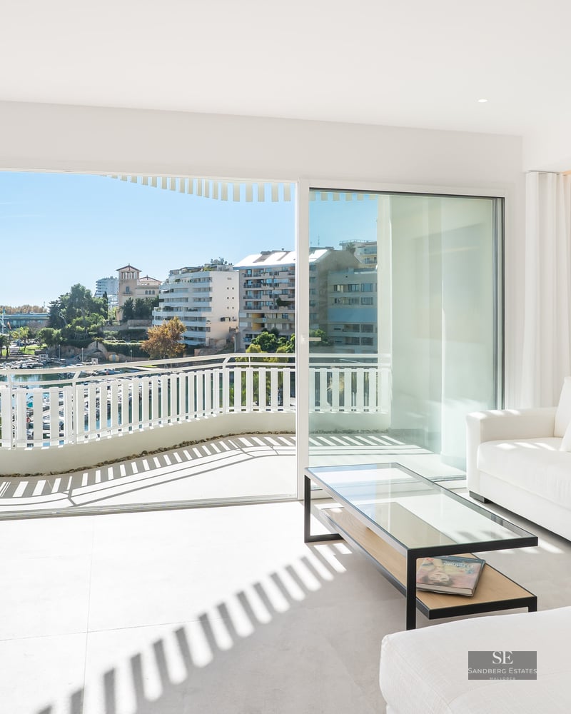 Bright living room with white sofas and glass doors opening to a balcony overlooking a marina.