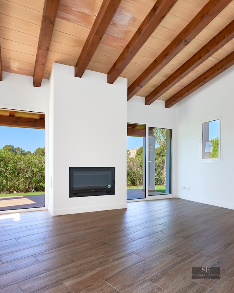 Spacious white living room with high ceiling, exposed wooden beams, fireplace, and sliding glass doors to a garden.