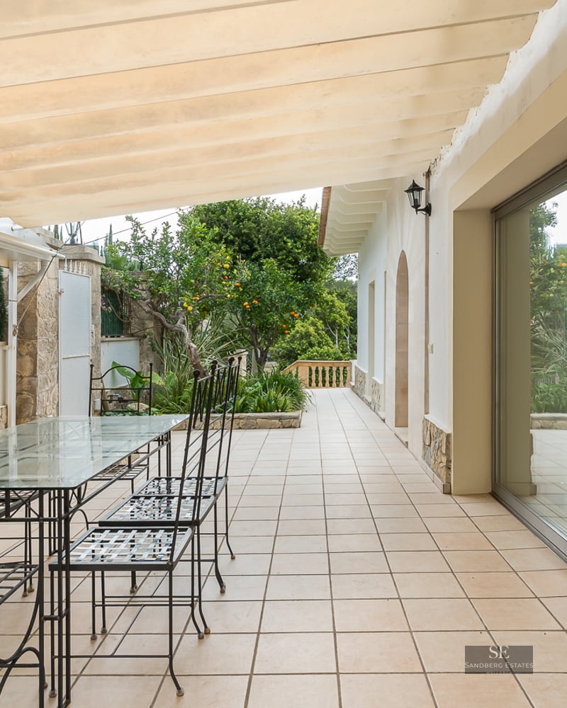 A shaded outdoor terrace with a glass dining table, black metal chairs, and a cream-colored retractable awning.