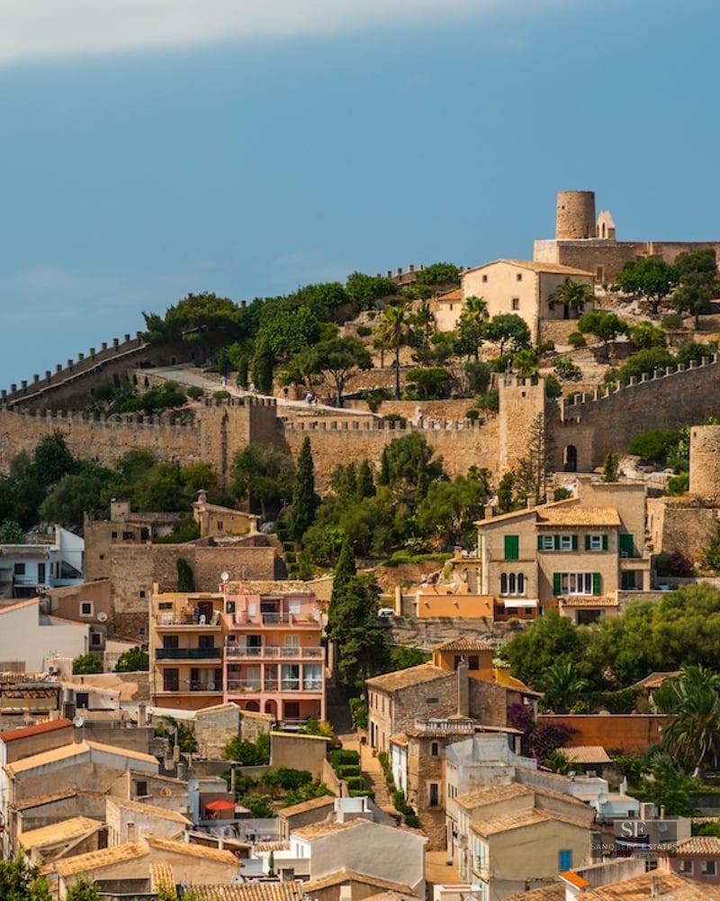 A large medieval stone castle with defensive walls on a lush green hill overlooking a historic town with tiled roofs.