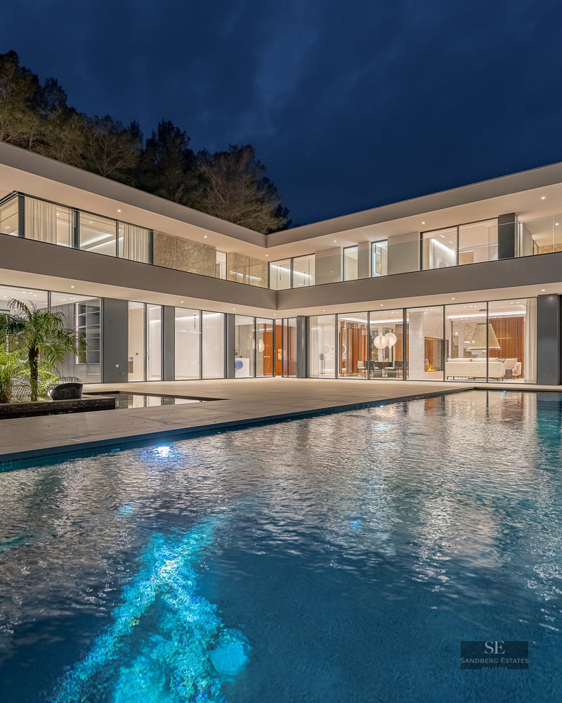 A luxury swimming pool in front of a glowing modern glass villa under a dark night sky.