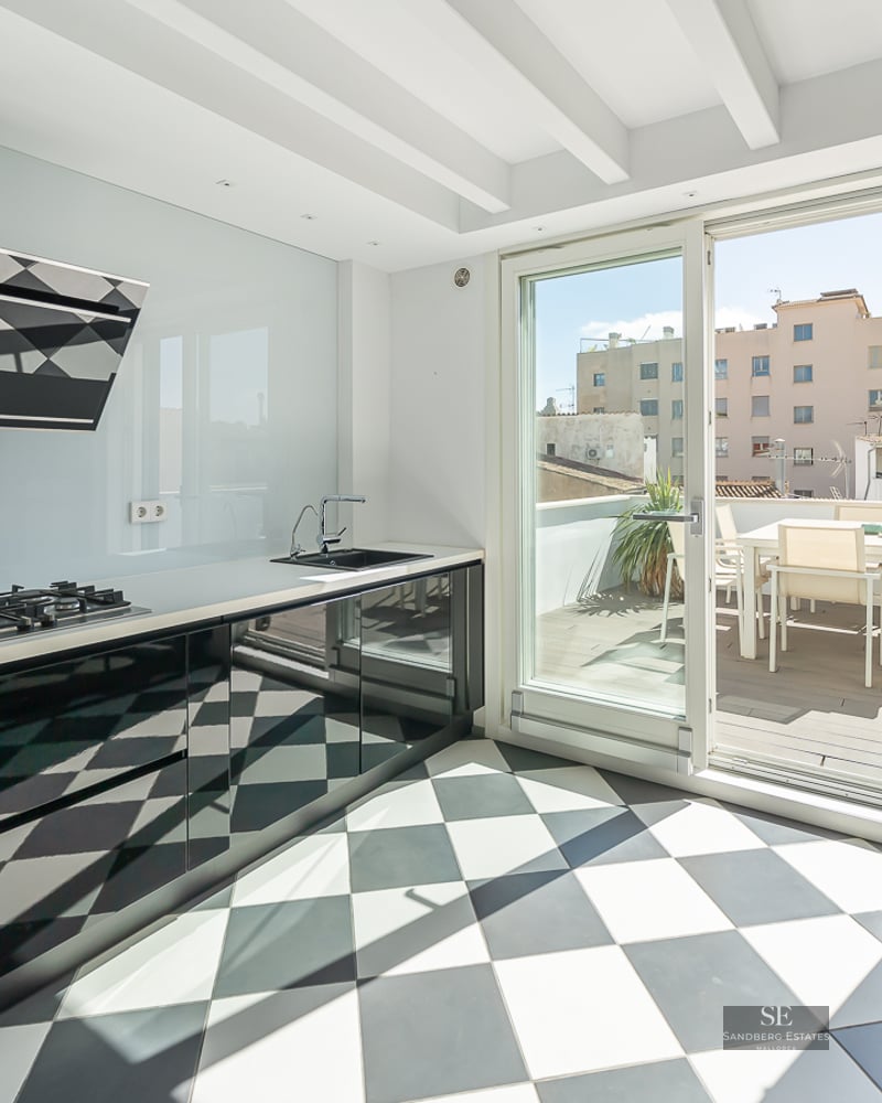 Modern kitchen with black and white checkered floor, glossy black cabinets, and a glass door leading to a sunny terrace.