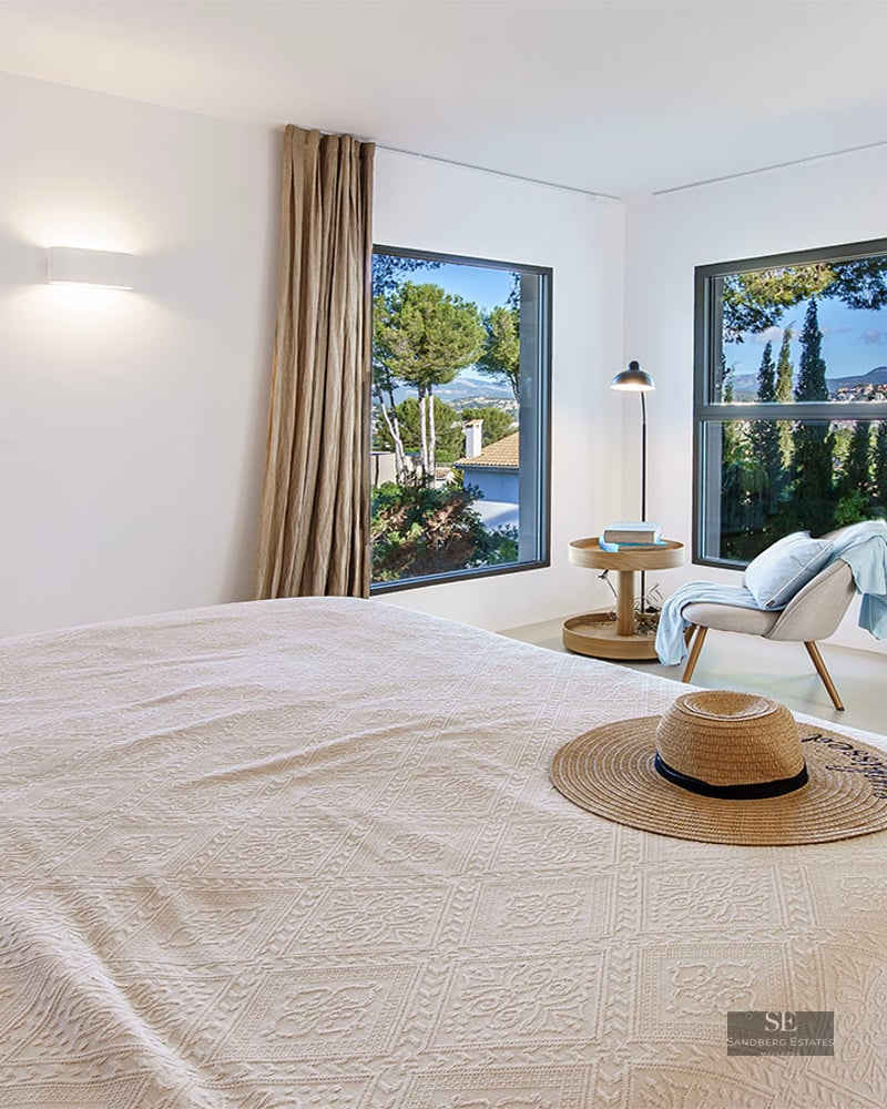 Modern bedroom with white walls, linen curtains, wooden furniture, and windows overlooking a lush green landscape.