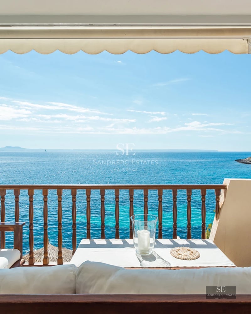 Sun-drenched Mediterranean terrace with white lounge sofas and wooden railing overlooking a sparkling turquoise sea.