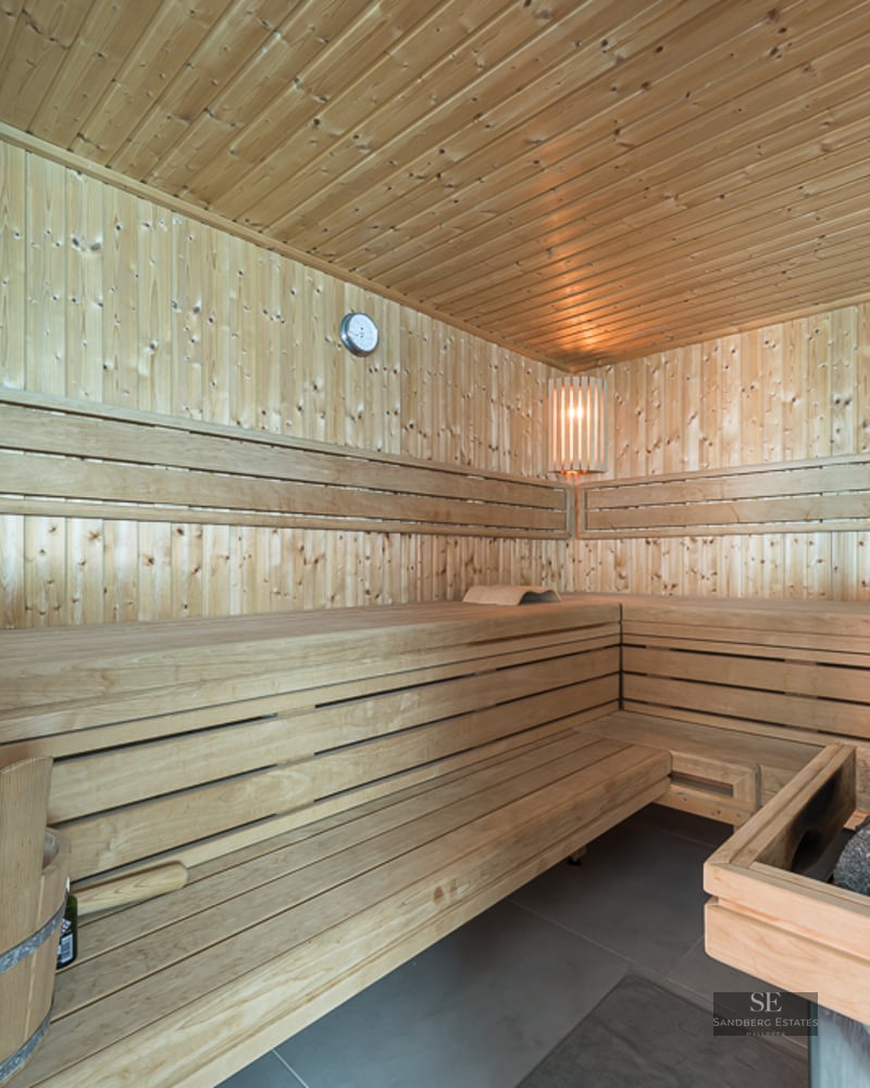 Interior of a modern wood-paneled sauna with tiered seating, a stone heater, and a wooden bucket.