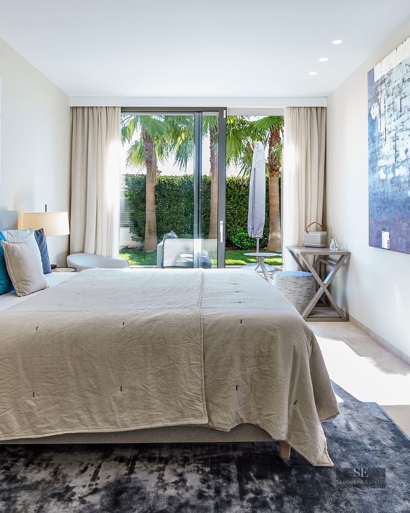 Chambre moderne avec grand lit, art abstrait et baies vitrées ouvrant sur un jardin avec des palmiers.