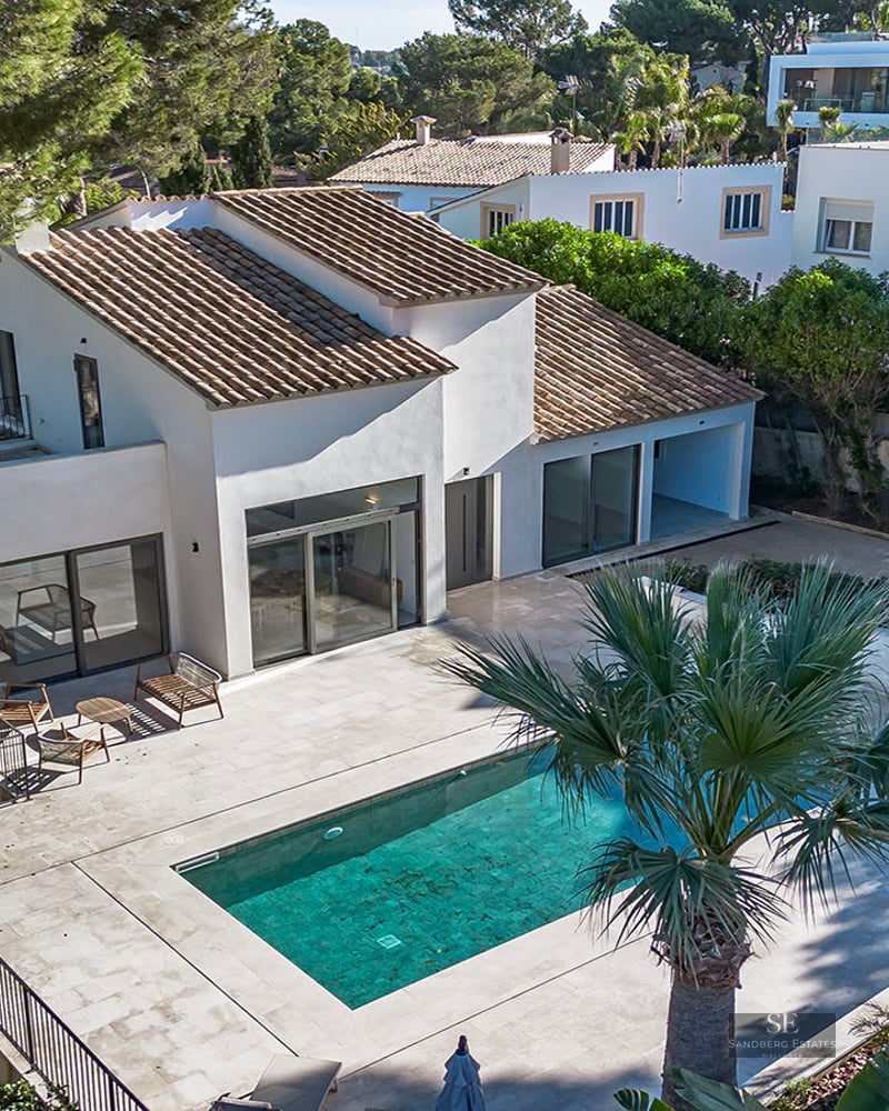 Aerial view of a white villa with a tiled roof, turquoise swimming pool, and large stone terrace surrounded by pine and palm trees.