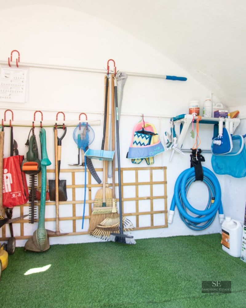 A white-walled storage room with artificial grass flooring containing a lawnmower, garden tools, and pool equipment.