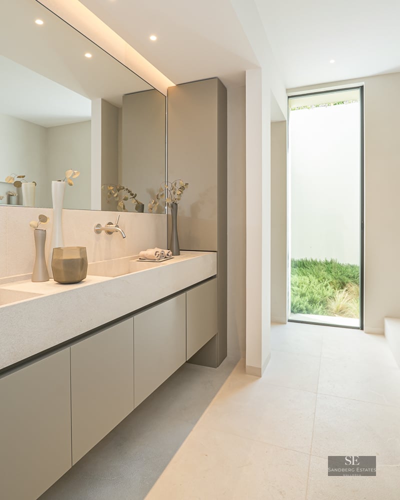 Minimalist bathroom with a stone double vanity, large mirror, freestanding white tub, and a window to a garden.