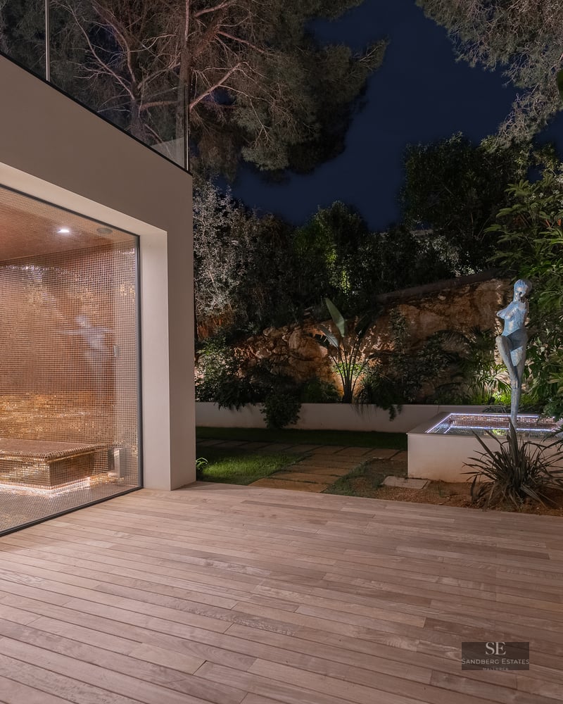 Glass-enclosed spa with gold mosaic tiles next to a wooden deck and illuminated garden with a sculpture at night.