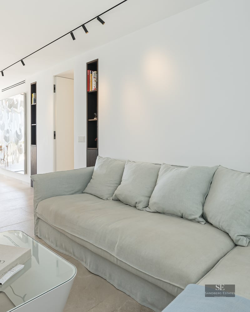 Bright living room featuring a large light grey sofa, glass coffee table, and dining area in the background with sea views.