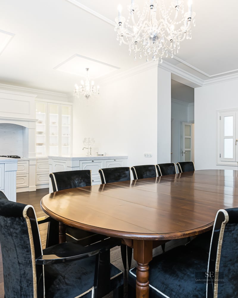 Large oval dark wood dining table surrounded by black velvet chairs under ornate crystal chandeliers in a white room.