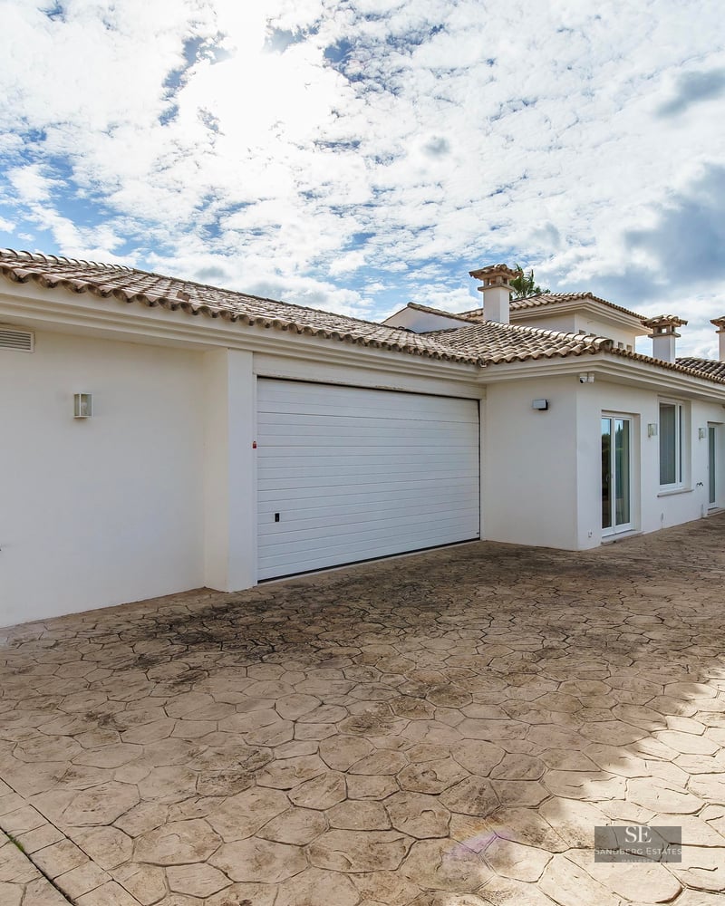 Spacious stamped concrete driveway with a white garage door in a Mediterranean style villa under a bright cloudy sky.