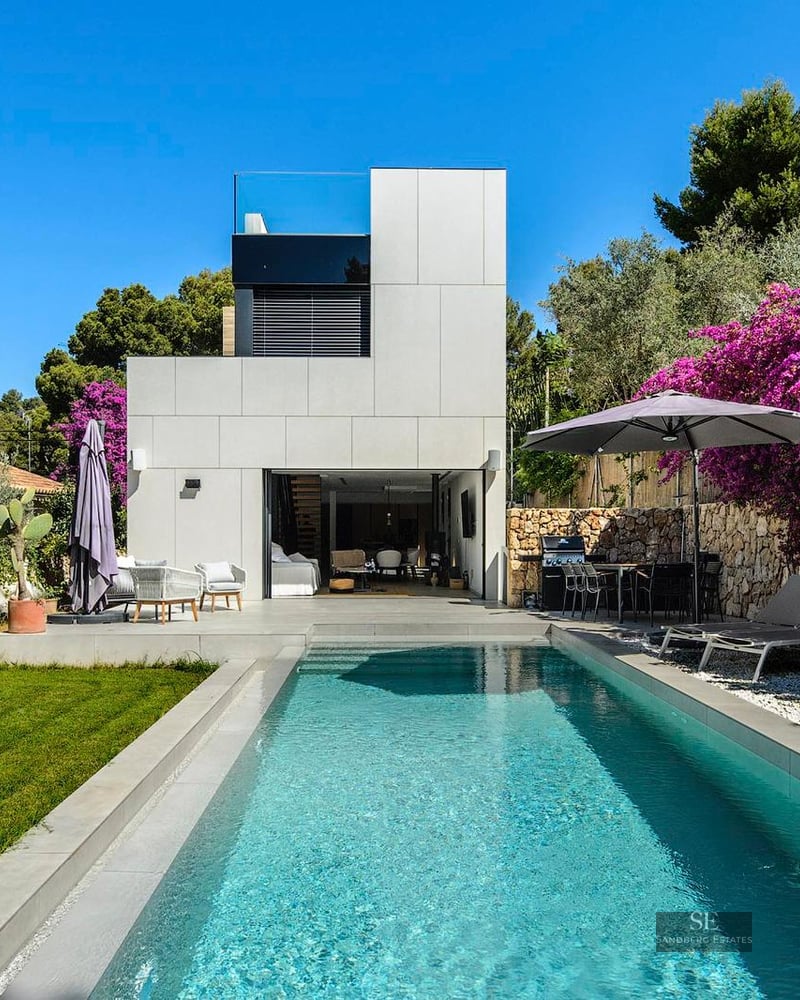 Modern white villa with a rectangular swimming pool, green lawn, and vibrant purple bougainvillea under a clear blue sky.