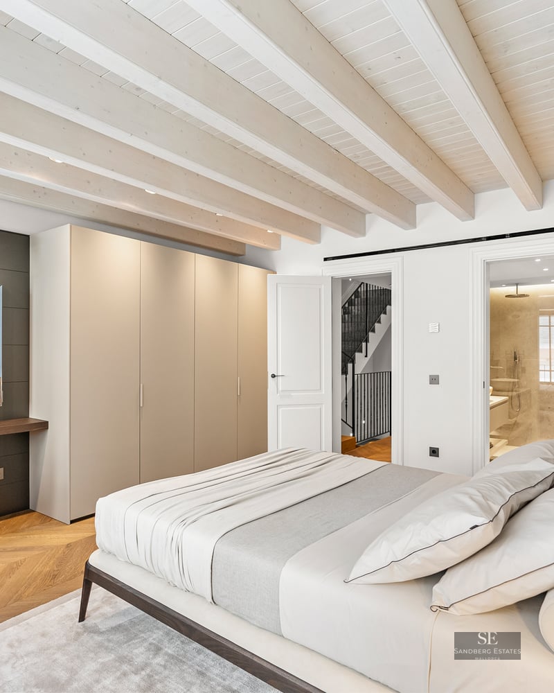 Luxury bedroom with white beamed ceiling, ensuite bathroom, built-in wardrobes, and wall-mounted TV over a desk.