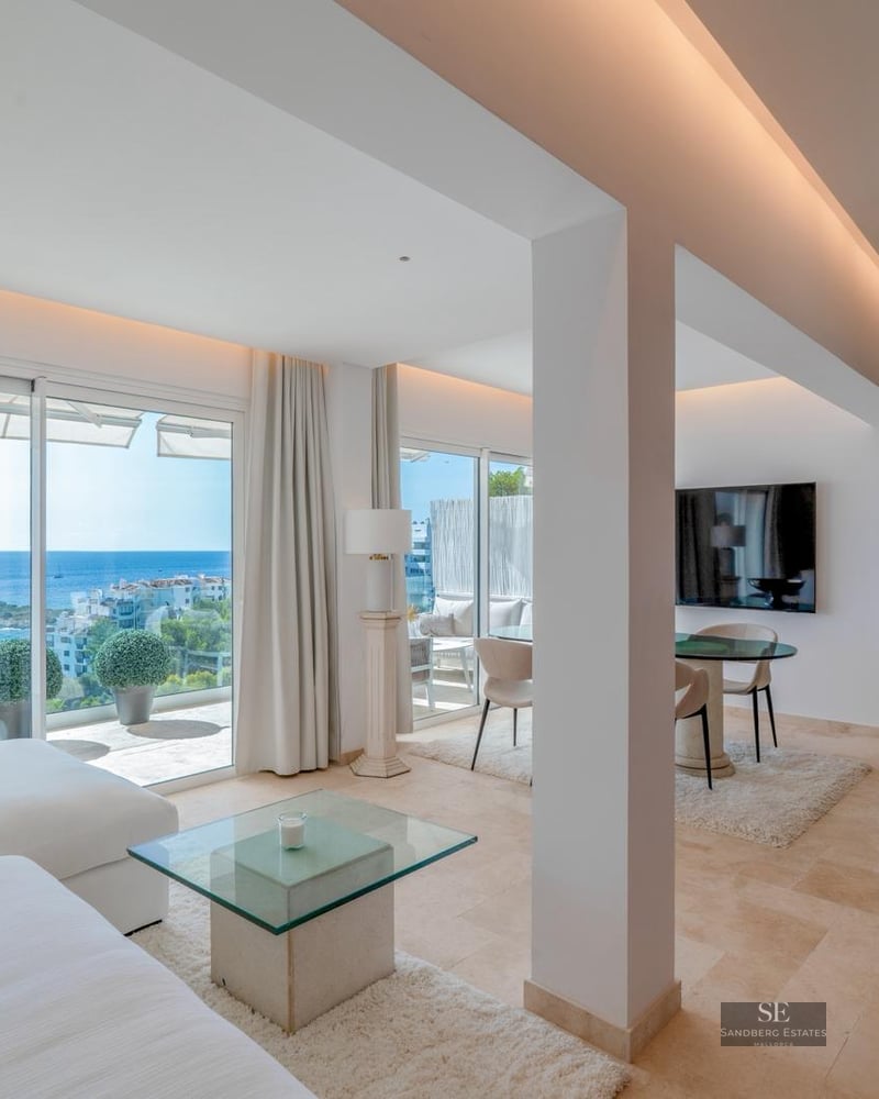 Bright modern living room with large glass doors opening to a Mediterranean sea view, featuring white sofas and stone floors.