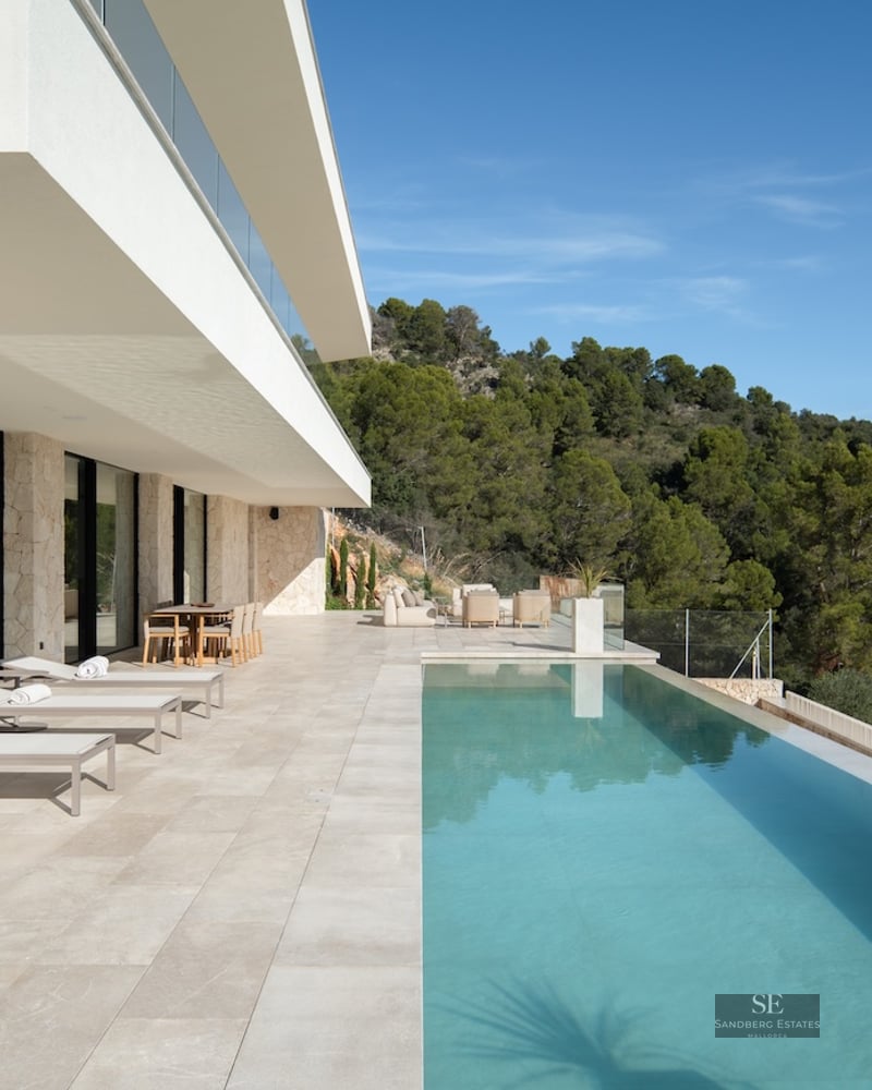 A spacious white stone terrace featuring an infinity pool, sun loungers, and views of a pine forest under a blue sky.