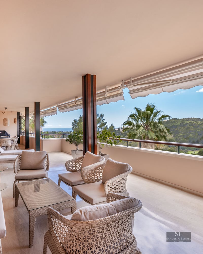 Grand balcon couvert avec mobilier en osier offrant une vue sur une vallée verdoyante et la mer par temps clair.