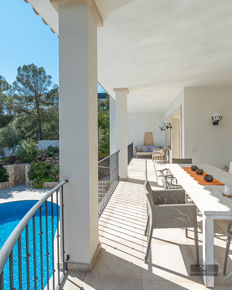 Terrasse couverte blanche avec grande table à manger et vue sur une piscine bleue et de la verdure.